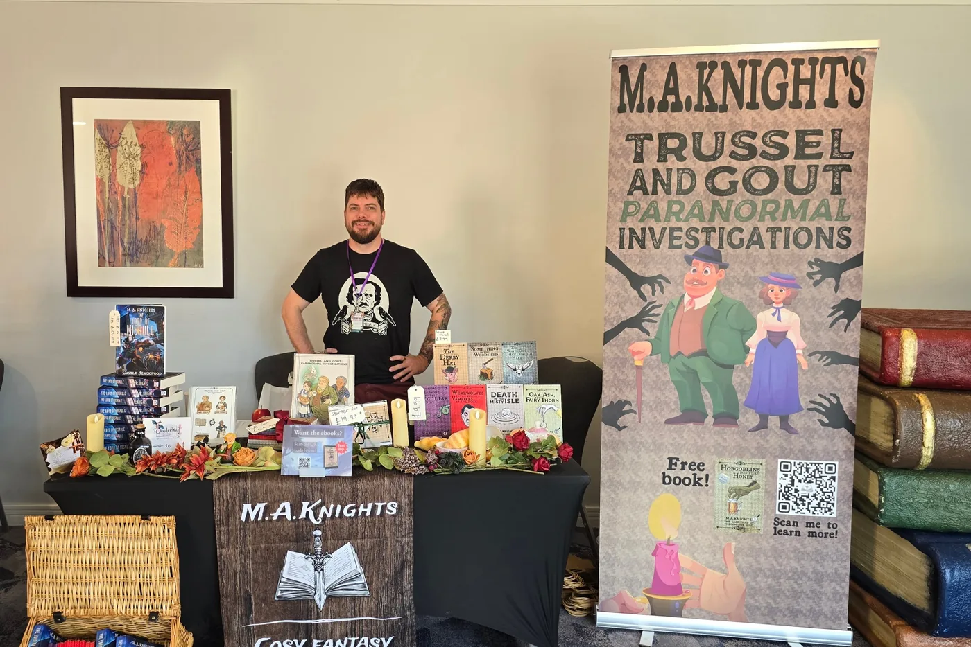 M.A. Knights standing behind his signing table: stacks of Trussel & Gout and The Lord of Misrule, candles and autumn flowers, with a tall Trussel & Gout banner beside it