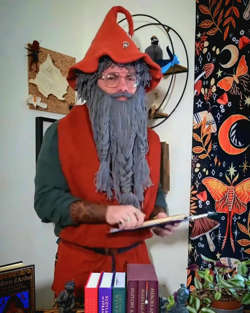 M.A. Knights dressed as Wilfred Bumblethump: red pointed hat, long grey plaited beard and round glasses, consulting a clipboard behind a stack of books